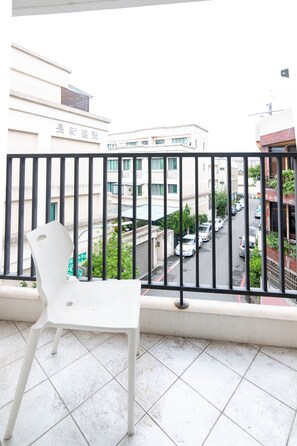 Un balcón con una silla de plástico blanco, piso de baldosas y vista a una calle con coches estacionados y edificios al fondo.
