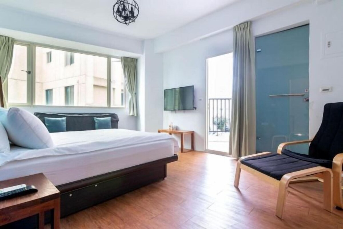 Ein modernes Hotelzimmer mit Bett, einem kleinen Tisch, einem Sessel, einem Fernseher und einem Balkon mit Vorhängen.