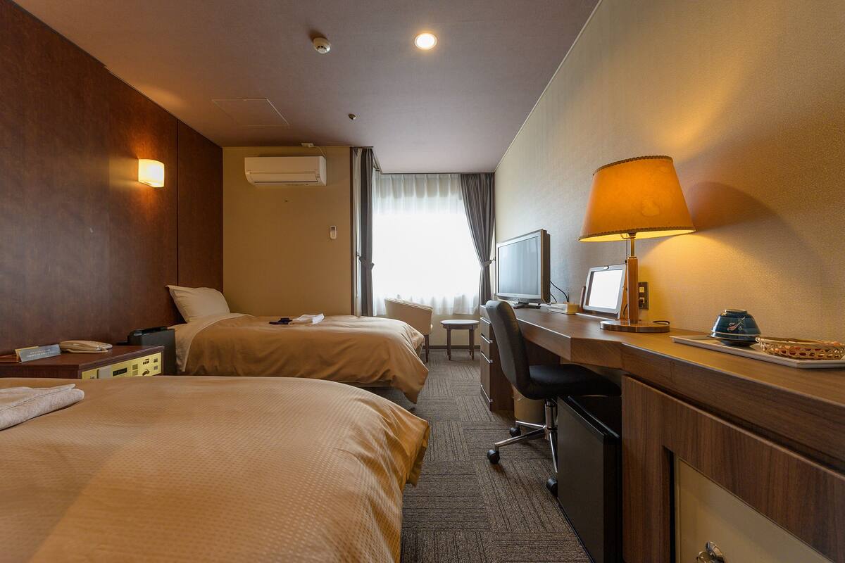 Ett hotellrum med två sängar, ett skrivbord med en dator, en lampa och ett fönster med gardiner.