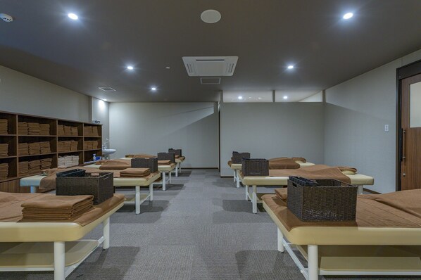 A room with multiple massage tables, each with a brown blanket and a basket. There are shelves with folded towels and a ceiling light.