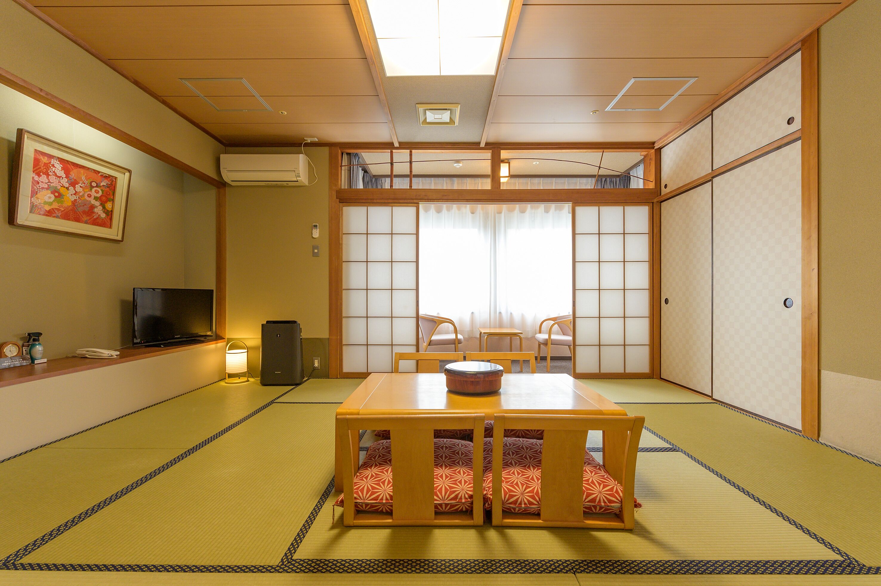 Ein traditioneller japanischer Raum mit Tatami-Boden, einem niedrigen Holztisch, zwei Stühlen, einem Fernseher und einer Schiebetür.