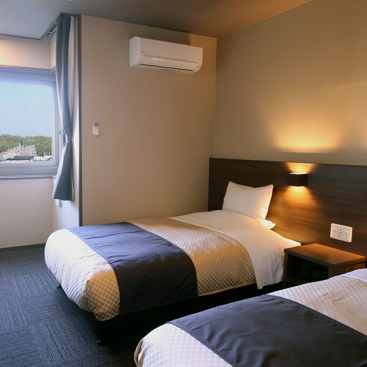 Ein Hotelzimmer mit zwei Betten, einer Klimaanlage, einem Nachttisch mit Lampe und einem Vorhangfenster.