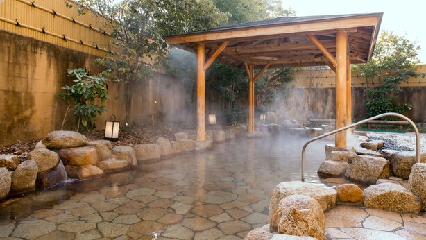 Een traditionele Japanse onsen met stenen vloeren, een houten paviljoen en een natuurlijke achtergrond.
