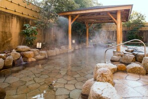 A traditional Japanese onsen with stone flooring, a wooden pavilion, and a natural backdrop.