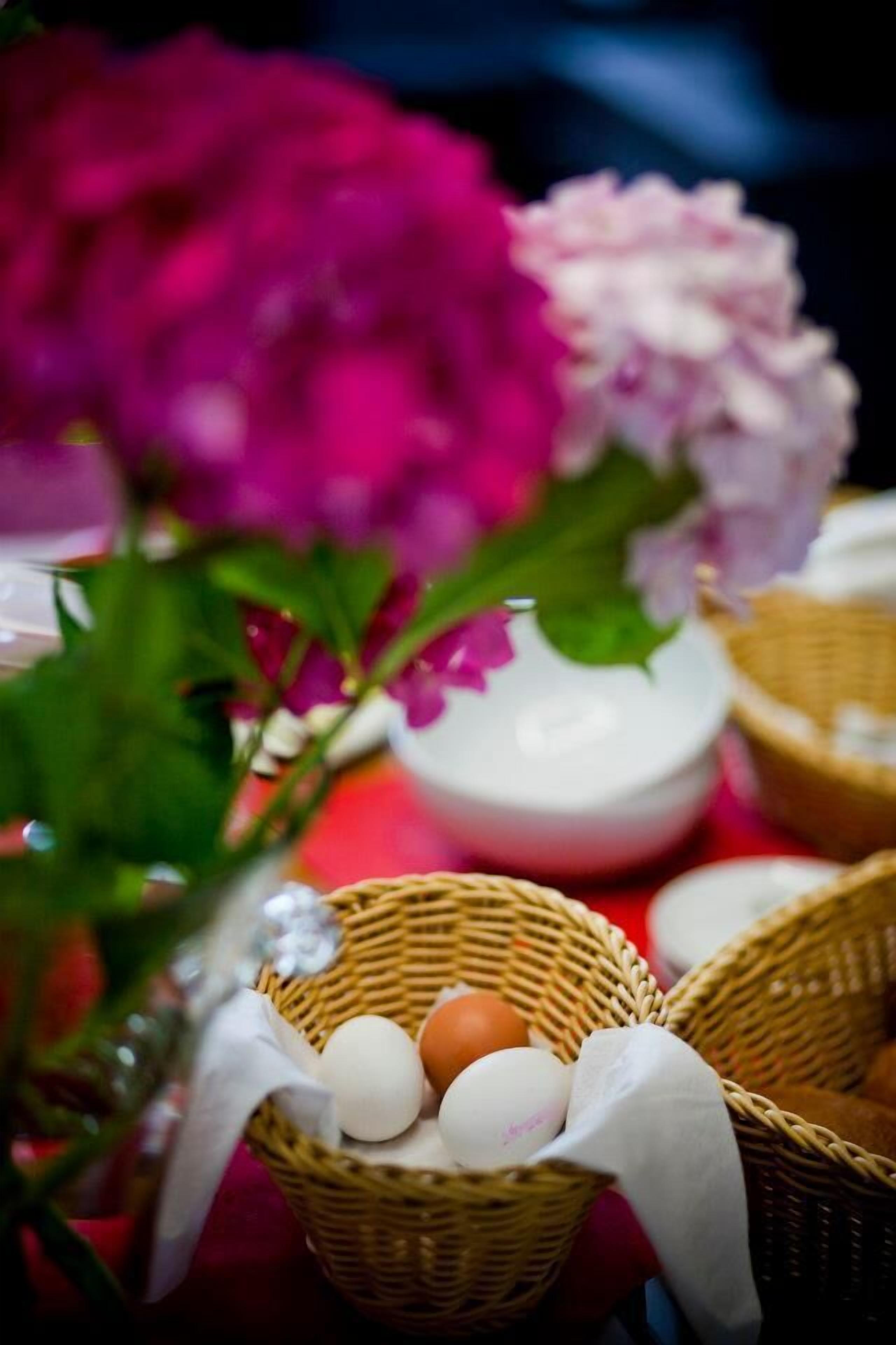 Primer plano de una cesta de mimbre con huevos y un paño blanco, sobre un fondo rojo con una flor rosa.