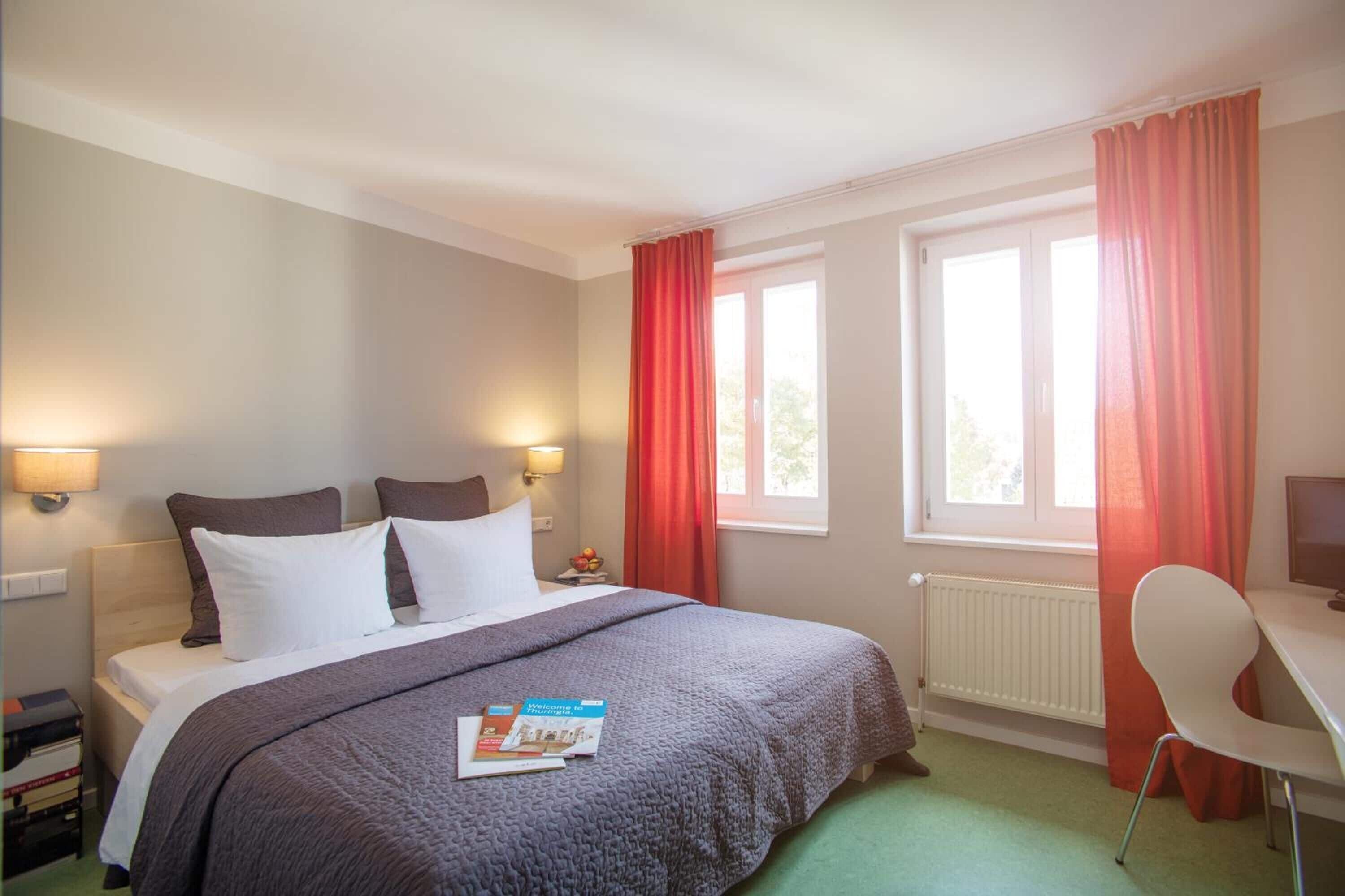 Una habitación de hotel con cama, dos lámparas de noche, un escritorio con silla y una ventana con cortinas.