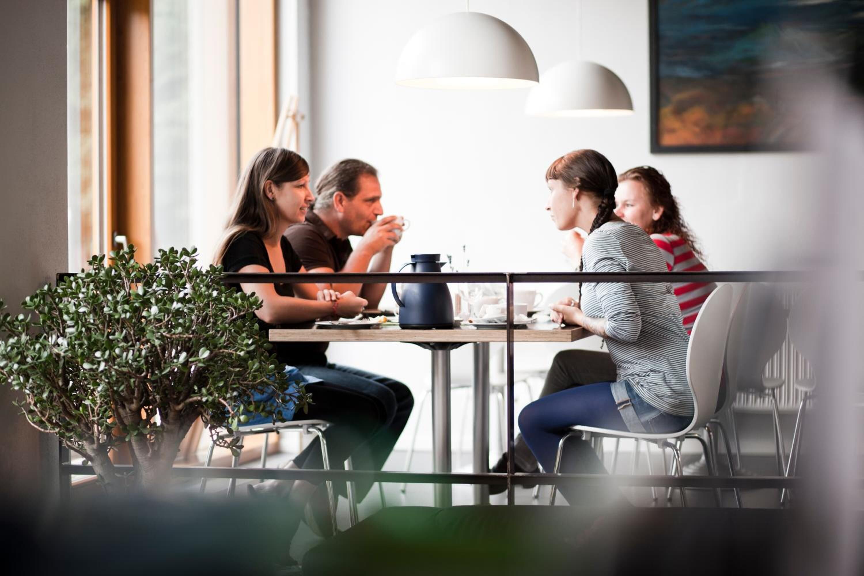 Un grupo de personas sentadas alrededor de una mesa en un café moderno con grandes ventanales y lámparas colgantes.
