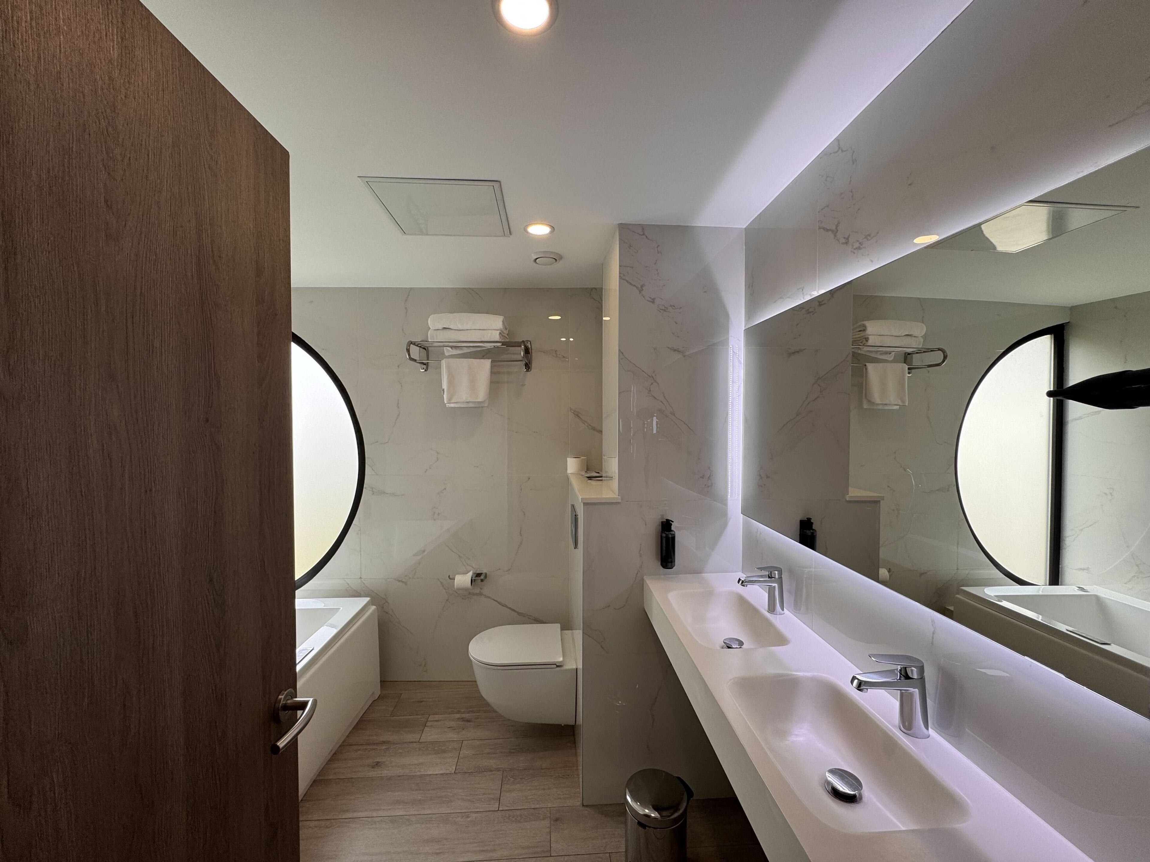 A modern bathroom with a large mirror, two sinks, a toilet, and a bathtub.