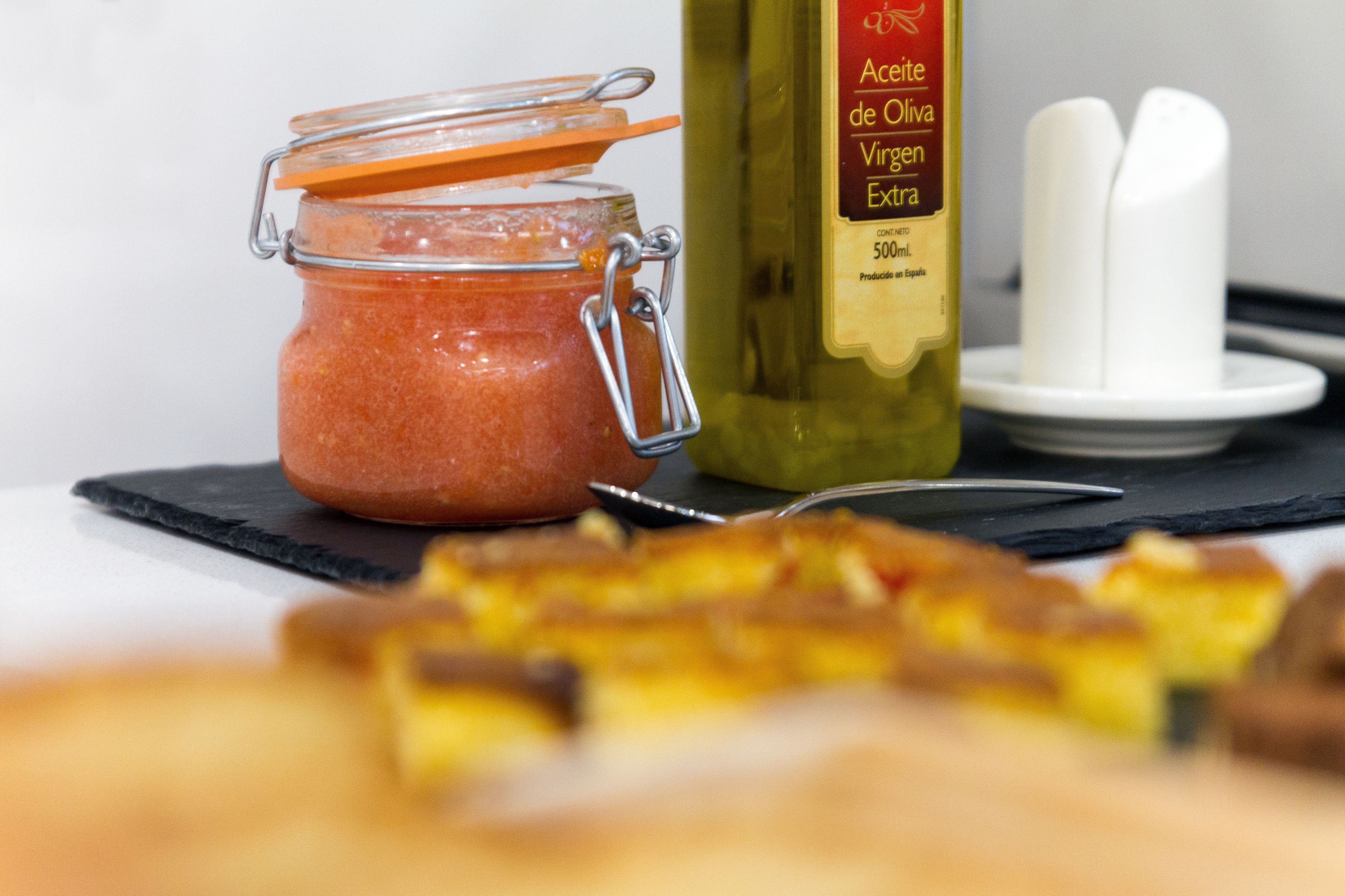 A jar of preserved olives, a bottle of olive oil, and a plate with a knife and spoon.