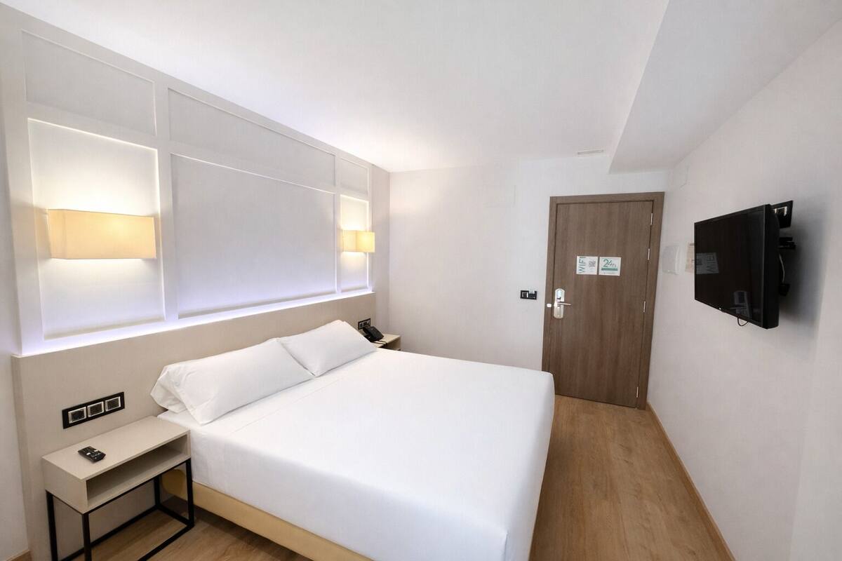A hotel room with a bed, bedside table, lamp, and a TV mounted on the wall.
