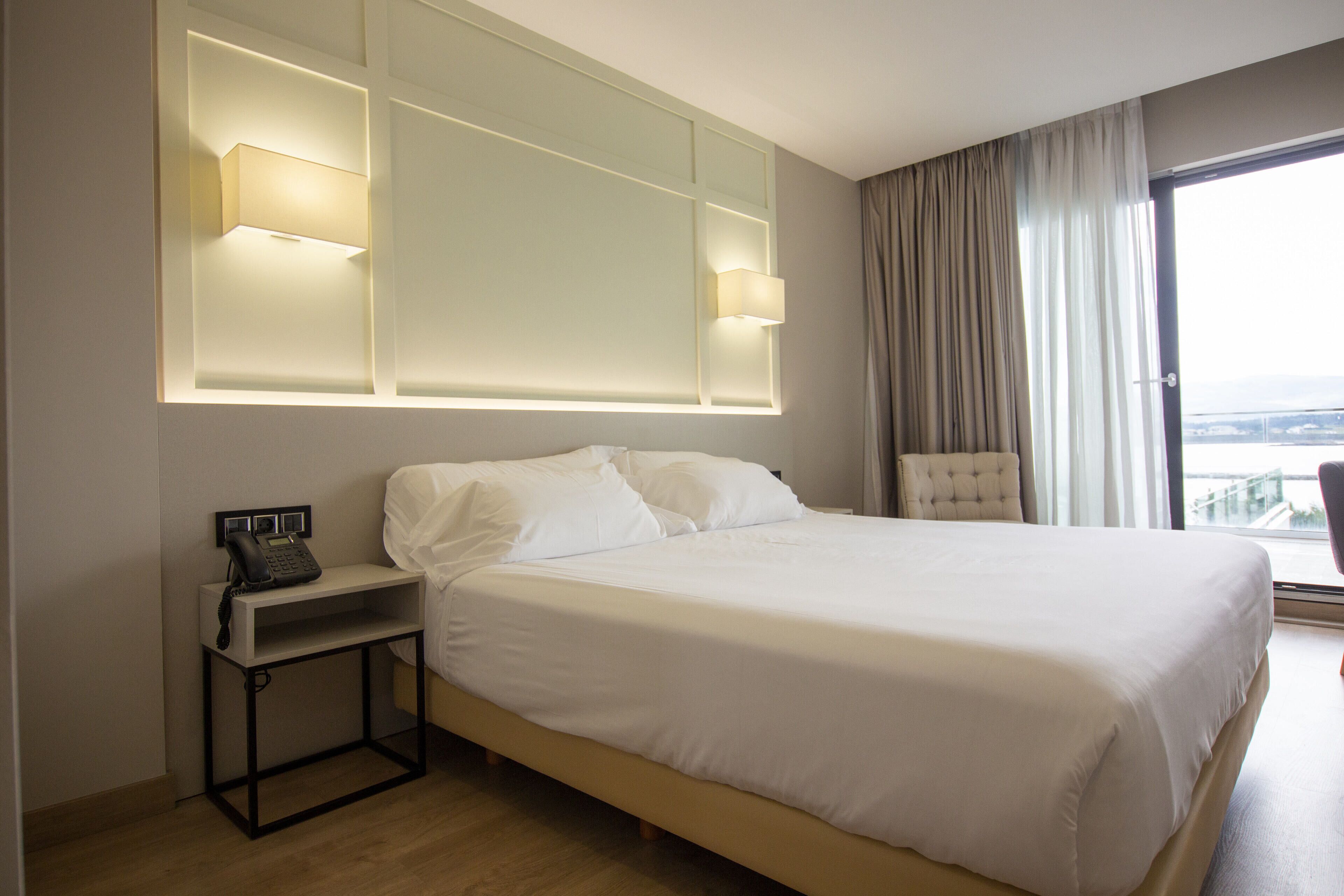 A hotel room with a large bed, bedside lamps, a telephone, and a view of the city through the window.