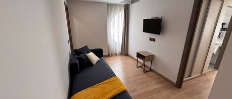 A modern hotel room with a dark sofa, a small wooden side table, a flat-screen TV mounted on the wall, and a sliding door leading to a bathroom.