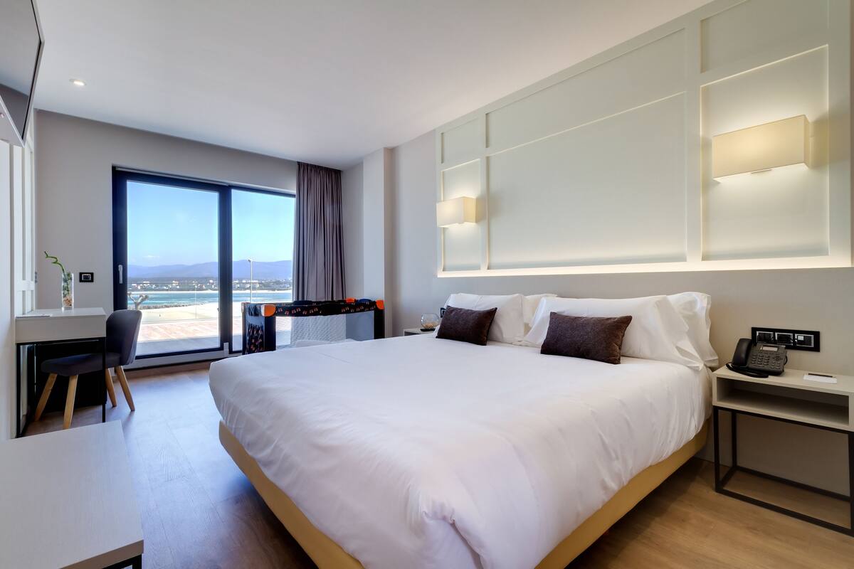 A modern hotel room with a large bed, a desk, a chair, and a view of the beach through the window.