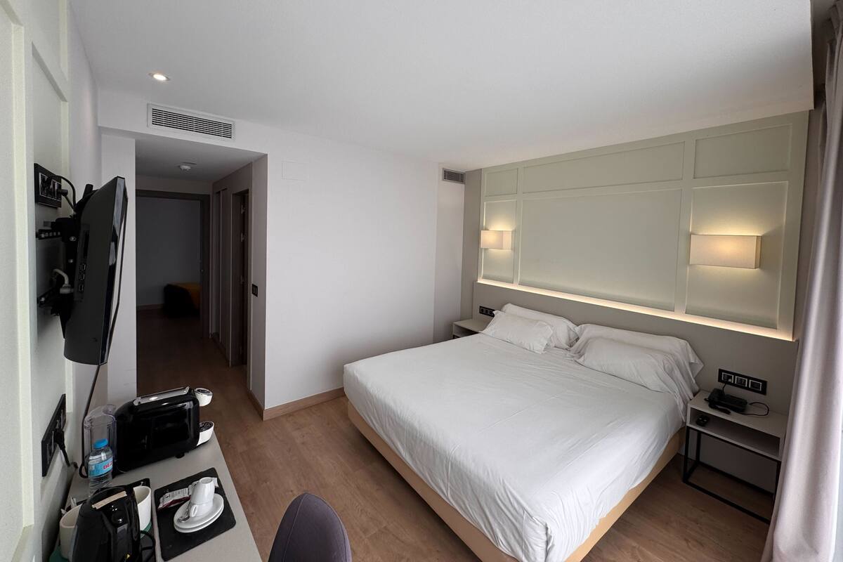 A hotel room with a bed, bedside table, lamp, and a desk with a computer and other items.