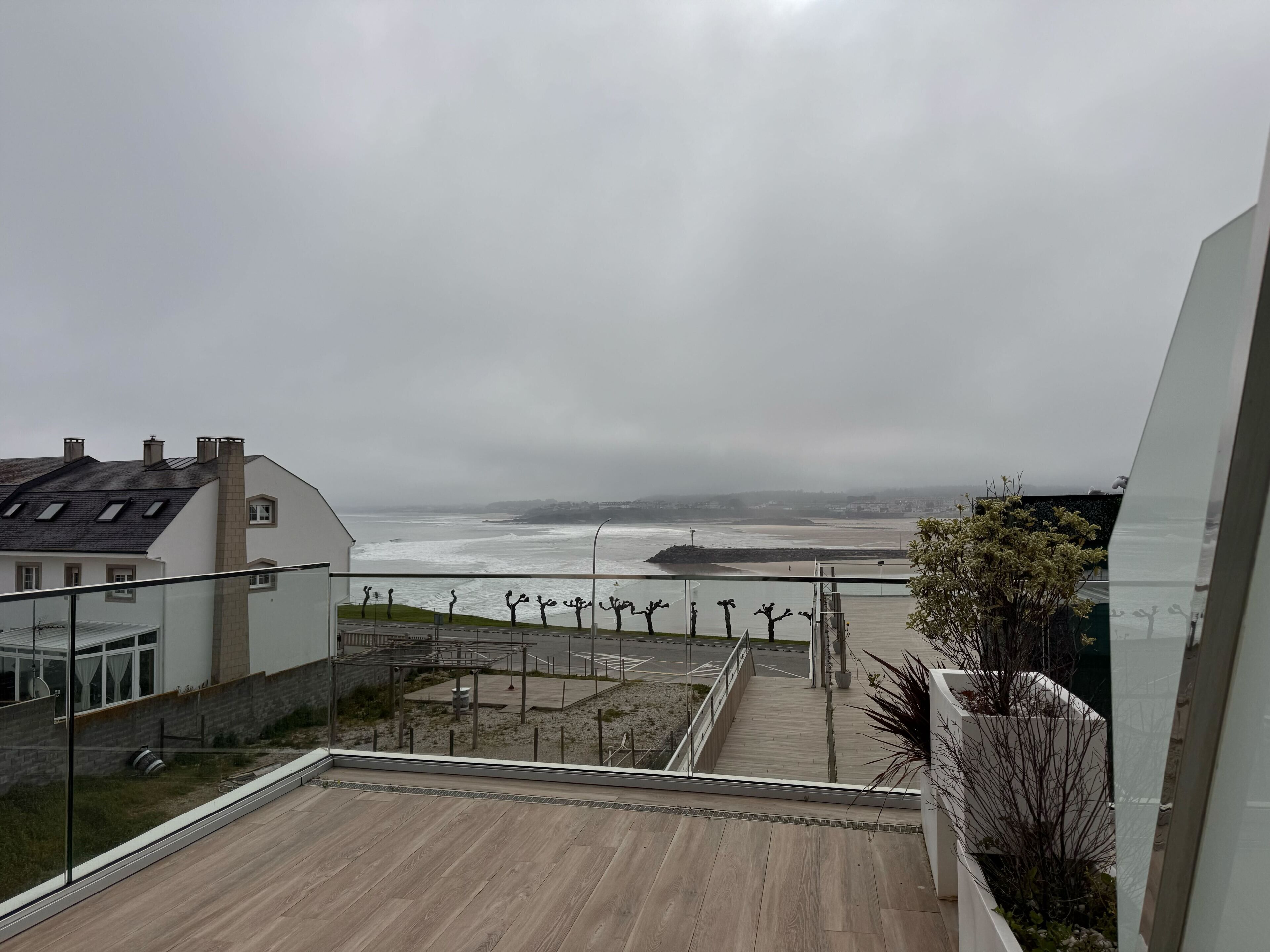 A coastal view from a balcony with a wooden floor, overlooking a beach and a cloudy sky.