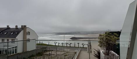 A coastal view from a balcony with a wooden floor, overlooking a beach and a cloudy sky.