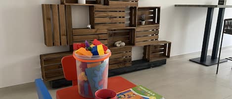 A room with a red table, a blue chair, a plastic container with colorful blocks, a red cup, and a book titled "PRIMERAS PINTURAS".