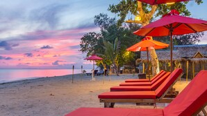 Ein Strand mit roten Liegestühlen und Sonnenschirmen, einer Struktur mit Reetdach und einem Sonnenuntergangshimmel.