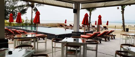 A poolside area with lounge chairs and tables, a pool, and a view of the beach.