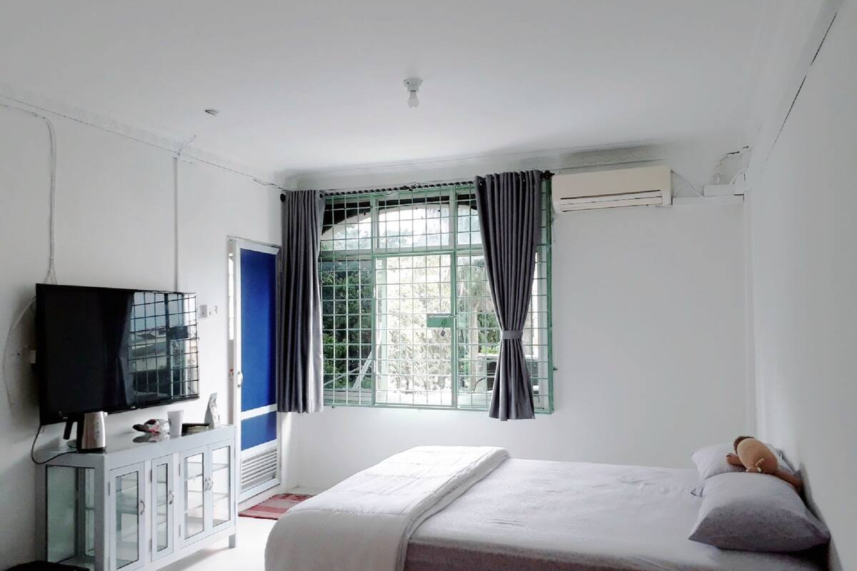 Ein Schlafzimmer mit einem Bett, einem Fernseher auf einem Schrank, einem Fenster mit Vorhängen und einer Klimaanlage.