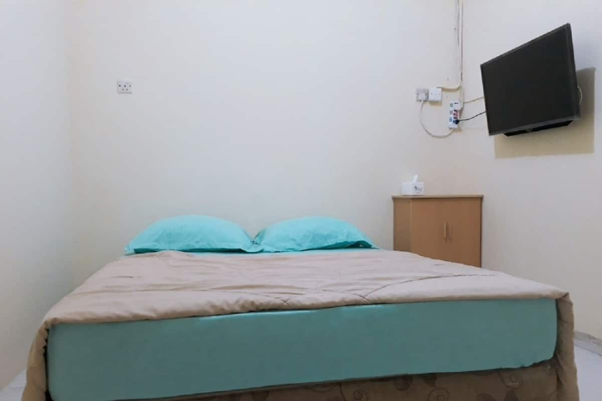Ein Einzelbett mit gemustertem Überwurf, ein kleiner Holzschrank und ein angebrachtes Fernsehgerät.