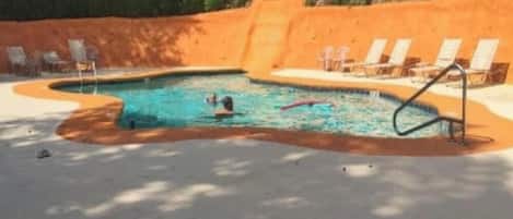 A swimming pool with a sandy border, surrounded by lounge chairs and a shaded area.