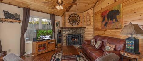 Interior acogedor de una cabaña con chimenea de piedra, sofá de cuero, mesa de centro de madera y televisor.