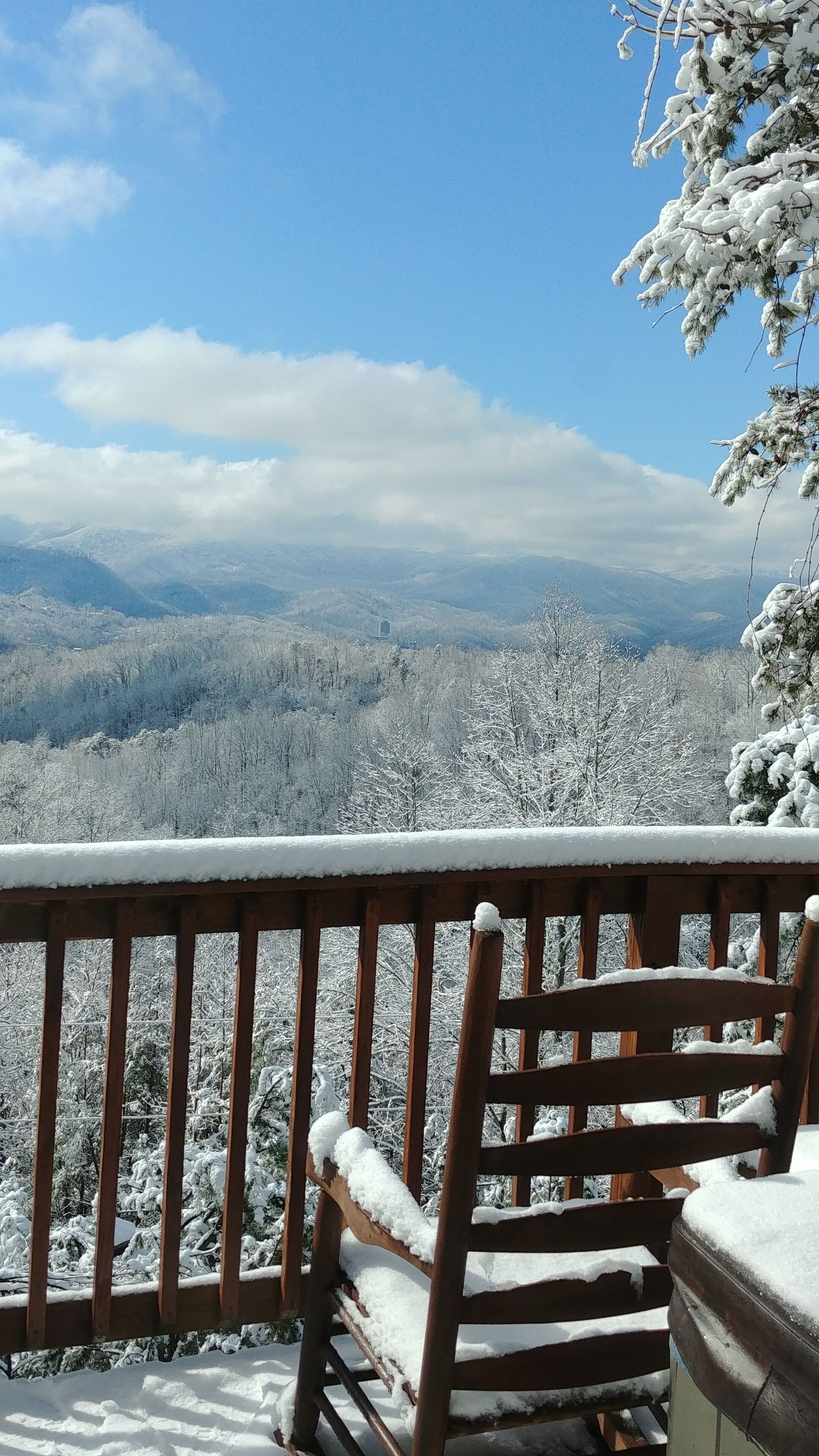 A wooden bench with snow on it, overlooking a snowy landscape with trees and mountains.