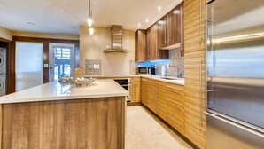 Une cuisine moderne dotée d’un grand îlot central, d’appareils électroménagers en acier inoxydable, de meubles de rangement en bois et offrant une vue sur l’extérieur.