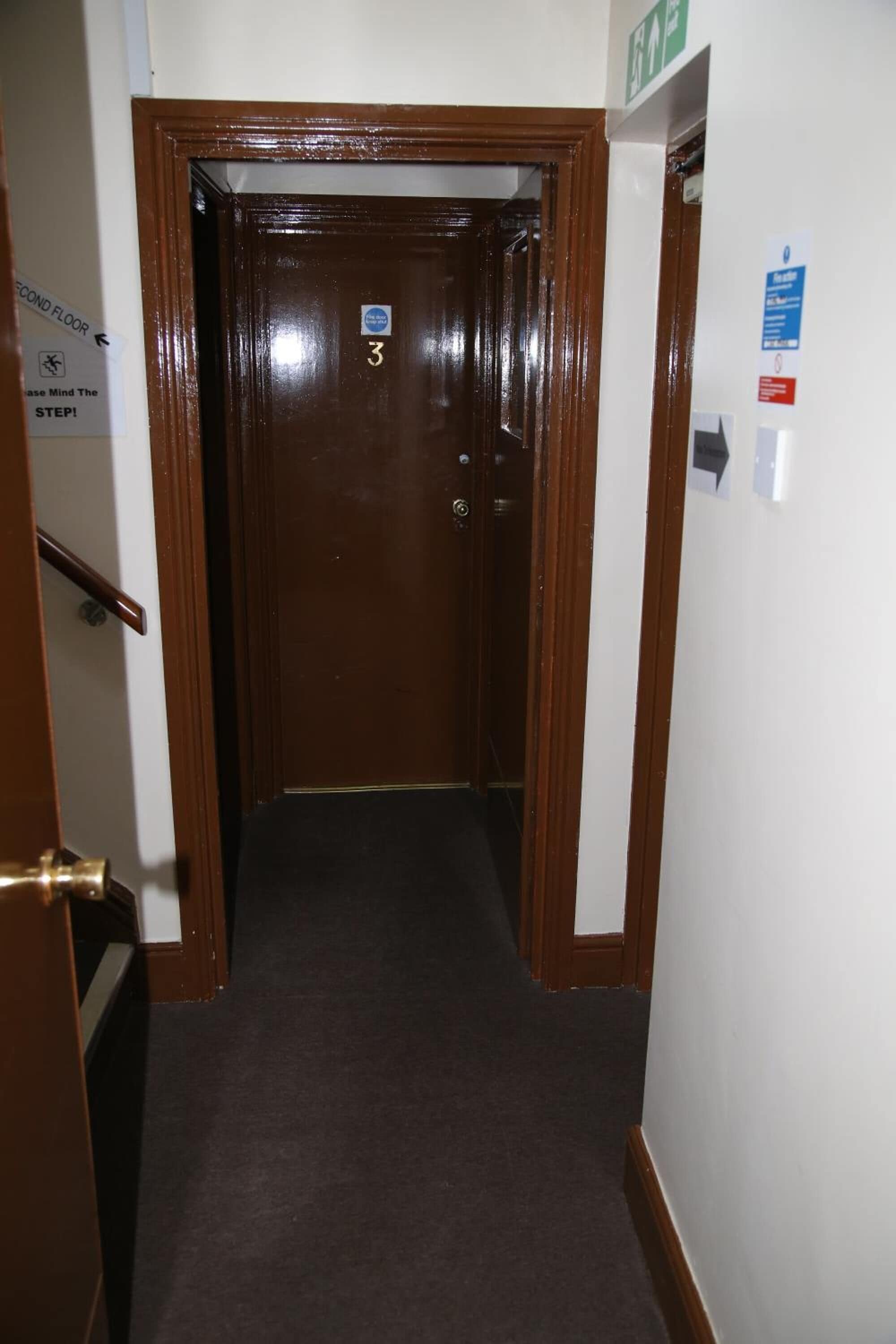 Un pasillo estrecho con una puerta de madera marcada con el número 3, una barandilla a la izquierda y un cartel que indica el segundo piso.