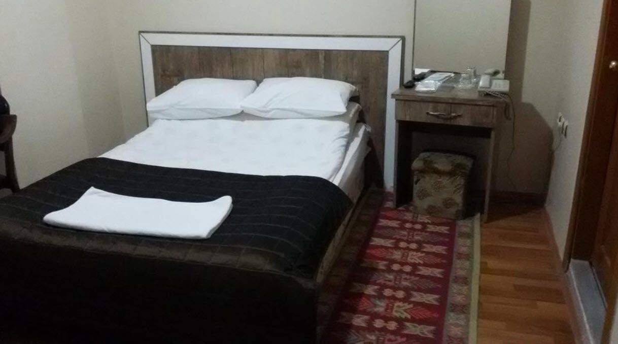 Una cama con colcha blanca y negra, un cabecero de madera, una mesita de noche con lámpara y una alfombra estampada en el suelo.