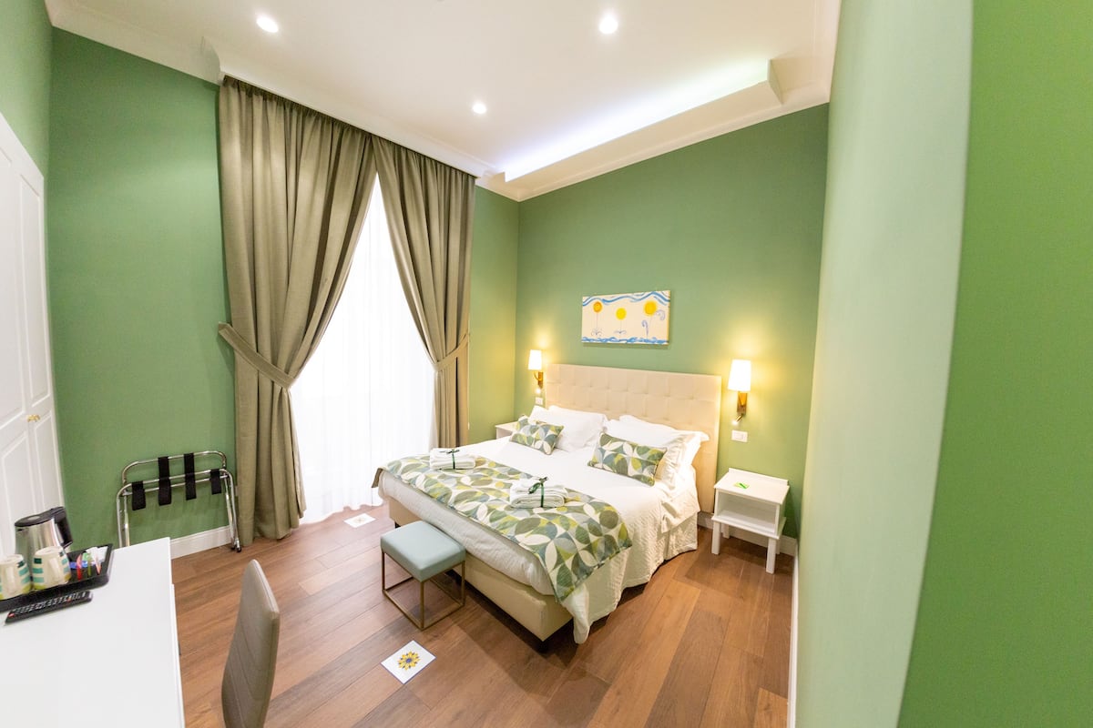 Habitación de hotel con cama, mesitas de noche, un banco, una silla, escritorio con bandeja de artículos de aseo y ventana con cortinas.