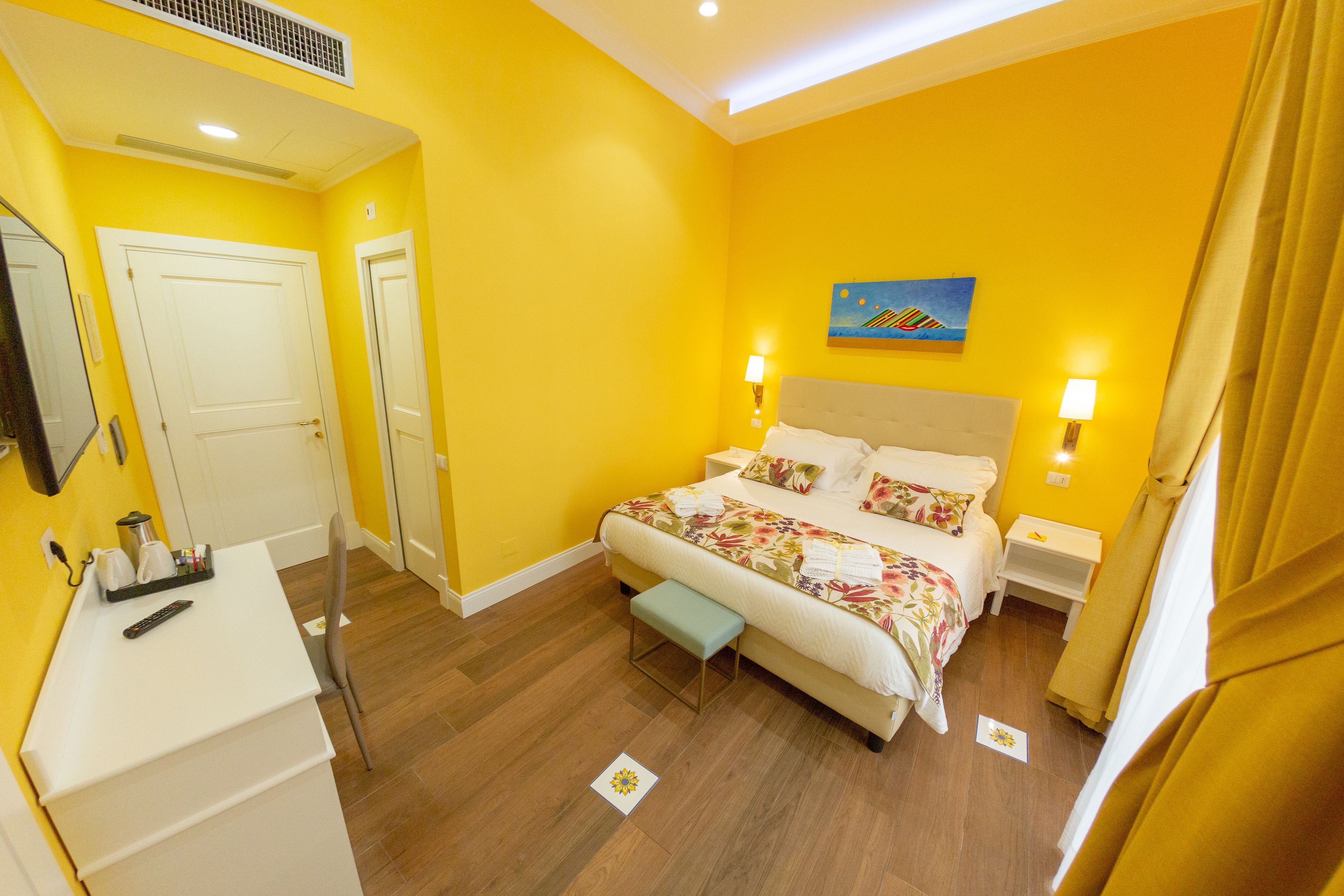 A hotel room with a bed, bedside tables, a desk with a chair, a mirror, and a painting on the wall.