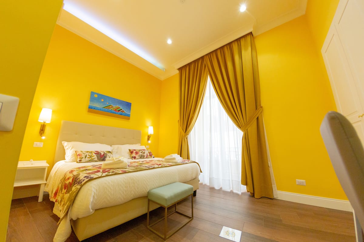 Una habitación de hotel con una cama grande, una mesita de noche, un banco y una ventana con cortinas.