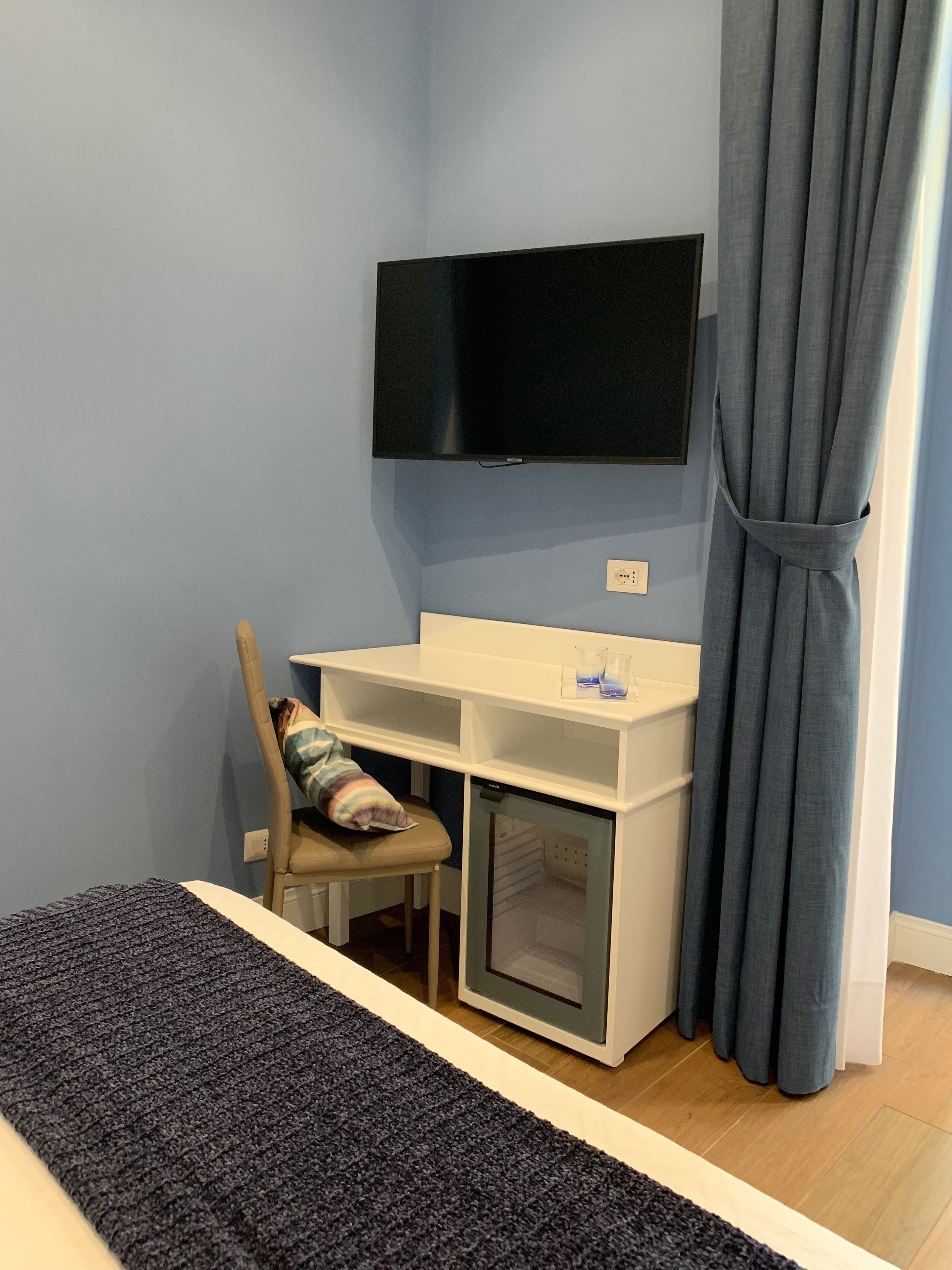 A room with a bed, a desk with a chair, a television mounted on the wall, and a window with curtains.