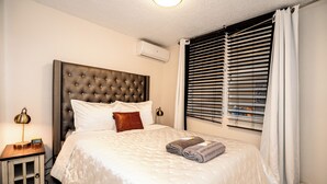 A neatly made bed with a tufted headboard, a bedside table with a lamp, and a window with blinds.