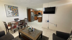 A living room with a dining area, a kitchen in the background, and a flat-screen TV mounted on the wall.