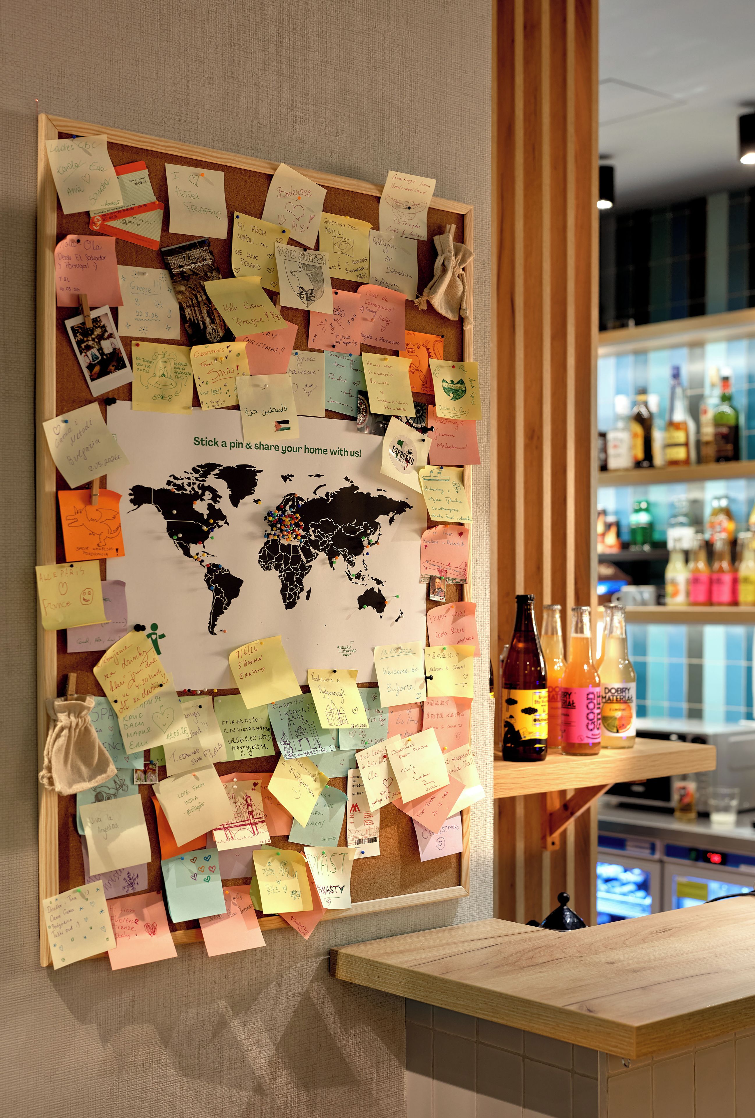 Un tablón de corcho con notas y un mapa del mundo, una barra de madera y una zona de bar con botellas.