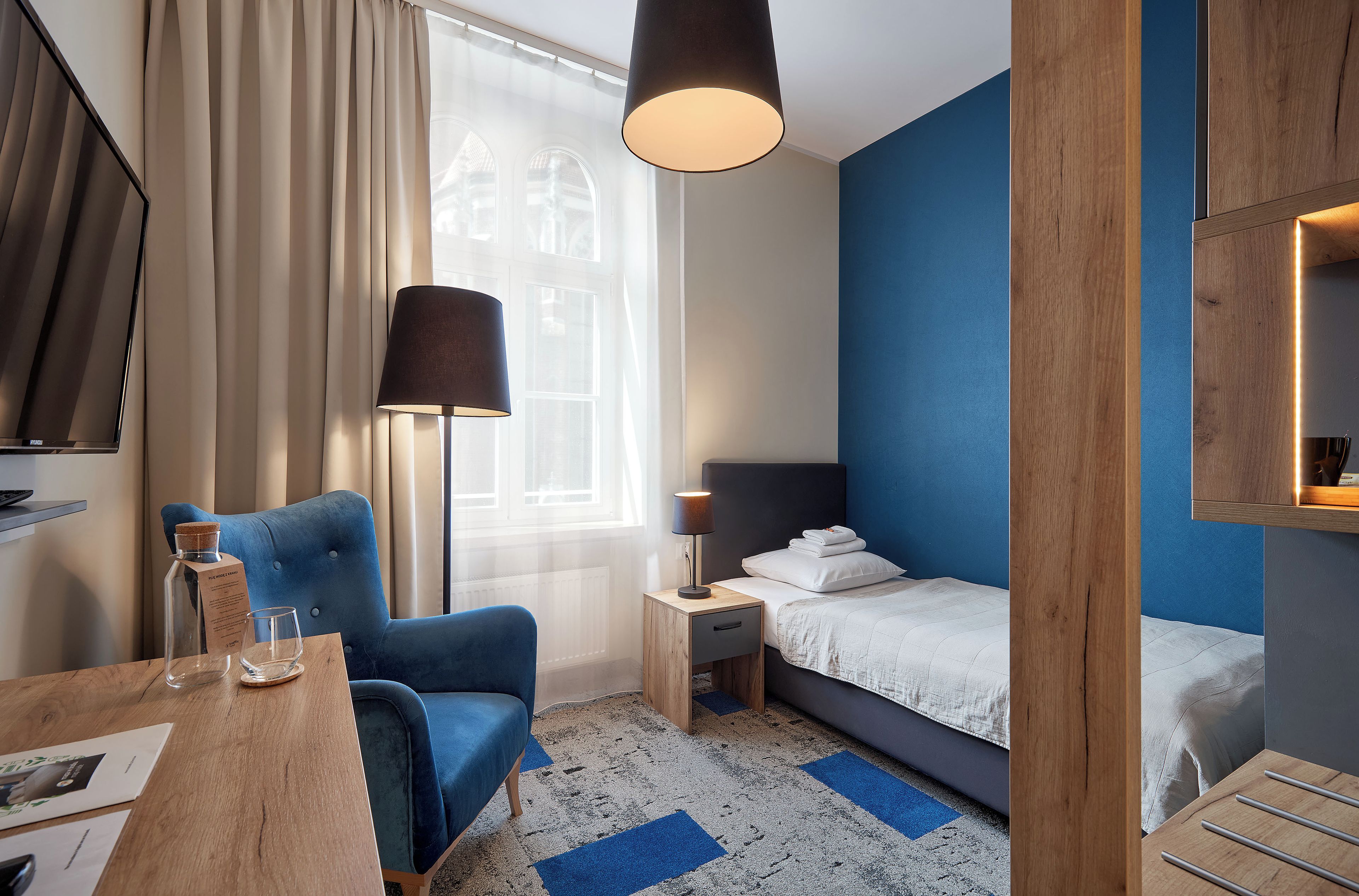 A hotel room with a bed, a chair, a table, a lamp, and a window with curtains.