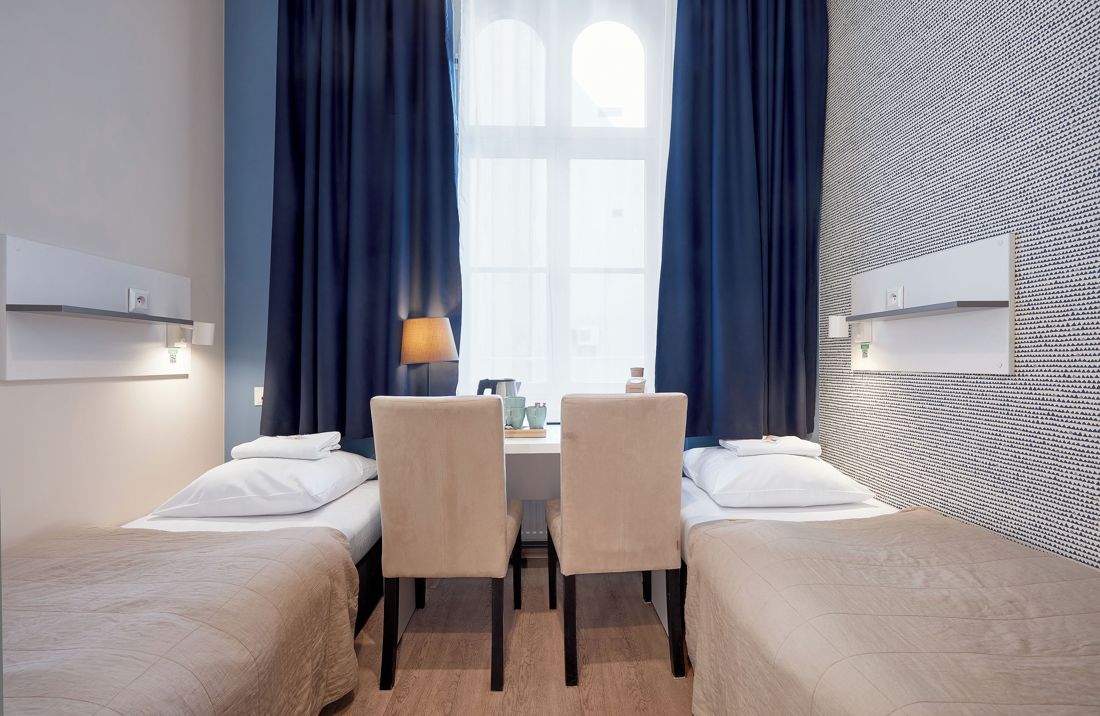 A hotel room with two single beds, a small table, chairs, and a window with curtains.