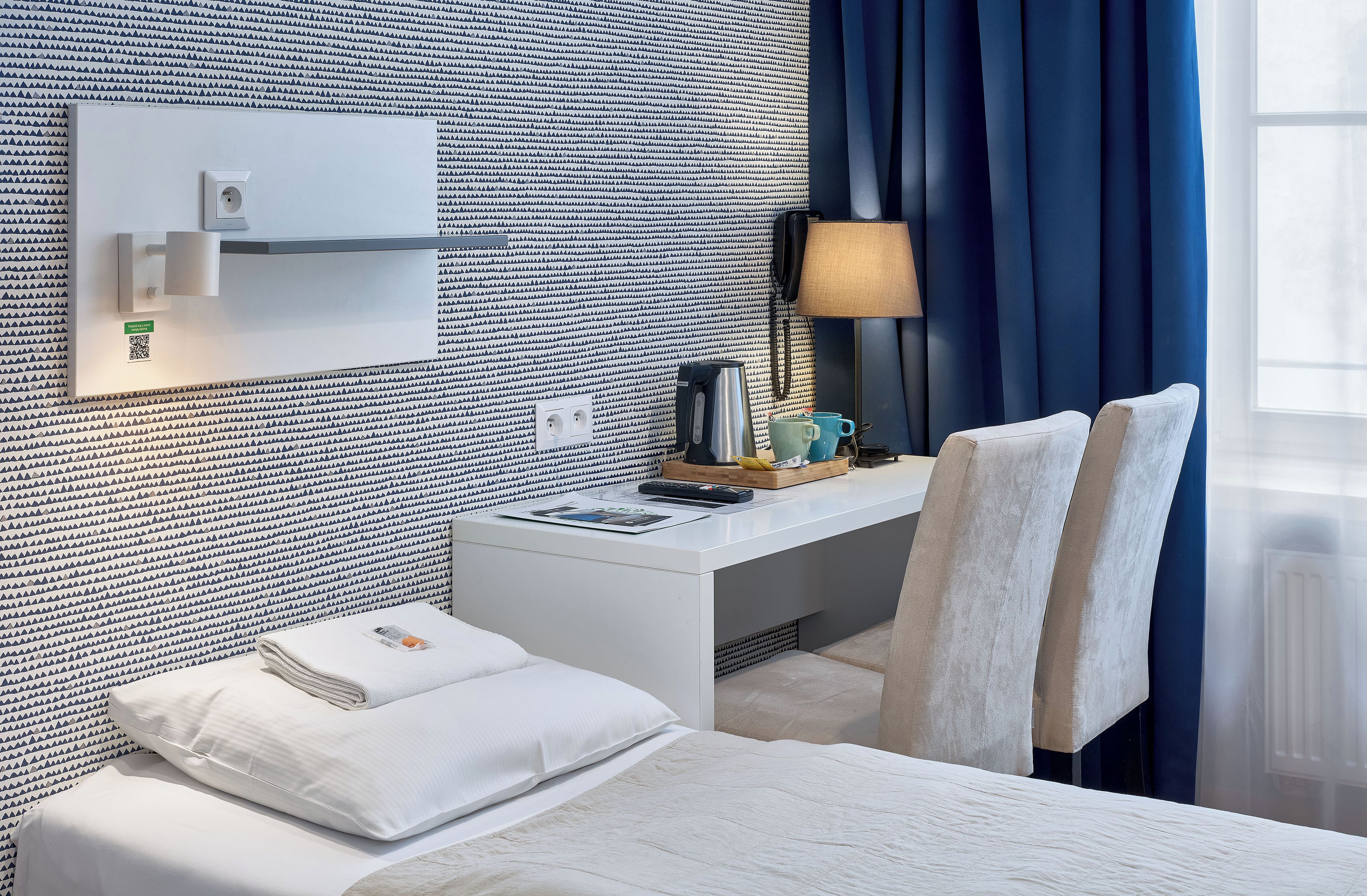 Quarto de hotel com cama, escrivaninha, luminária, cafeteira e janela com cortinas.