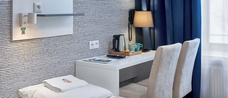 A hotel room with a bed, a desk with a lamp, a coffee maker, and a window with curtains.
