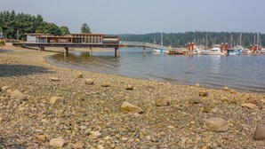 A riverside hotel with a wooden deck extending over the water, a bridge connecting to the shore, and boats docked at the marina. - Direct Ocean Front Access/private entry I BDRM, full bathroom, all season patio. (Mill Bay)