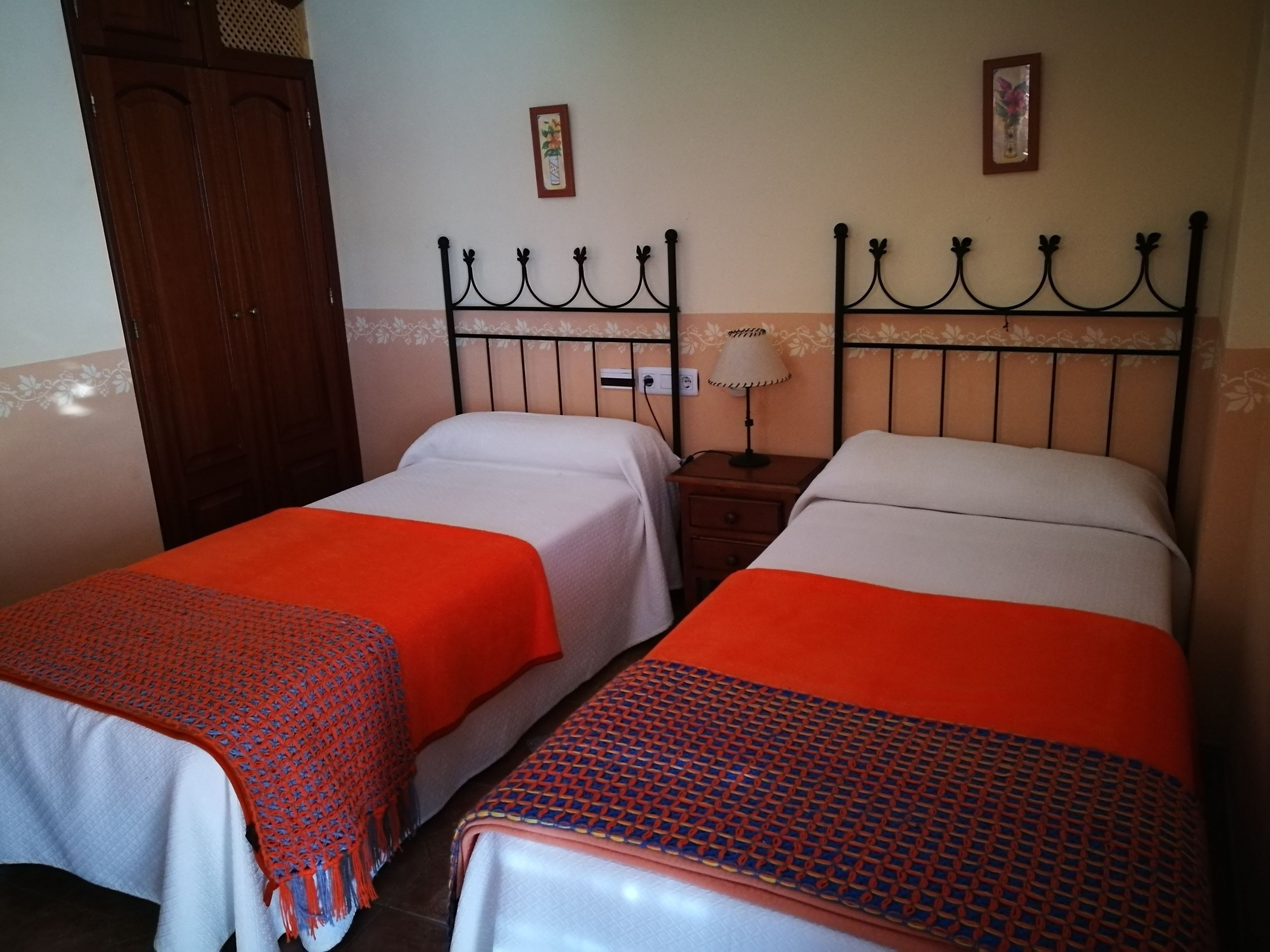 A room with two single beds, each with a red and white patterned bedspread. There is a wooden wardrobe to the left and a nightstand with a lamp between the beds.