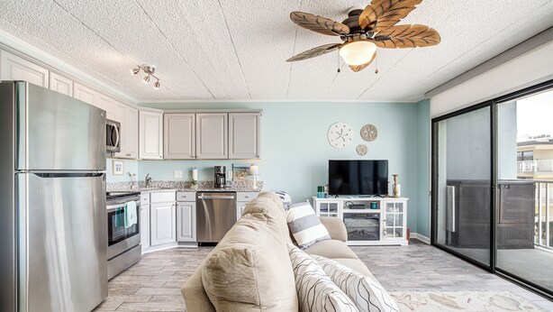 A spacious living room with a beige sofa, a ceiling fan, and a kitchen area with white cabinets and stainless steel appliances. - Stroll to Shore! Condo w/ Balcony in Ocean City (Ocean City)