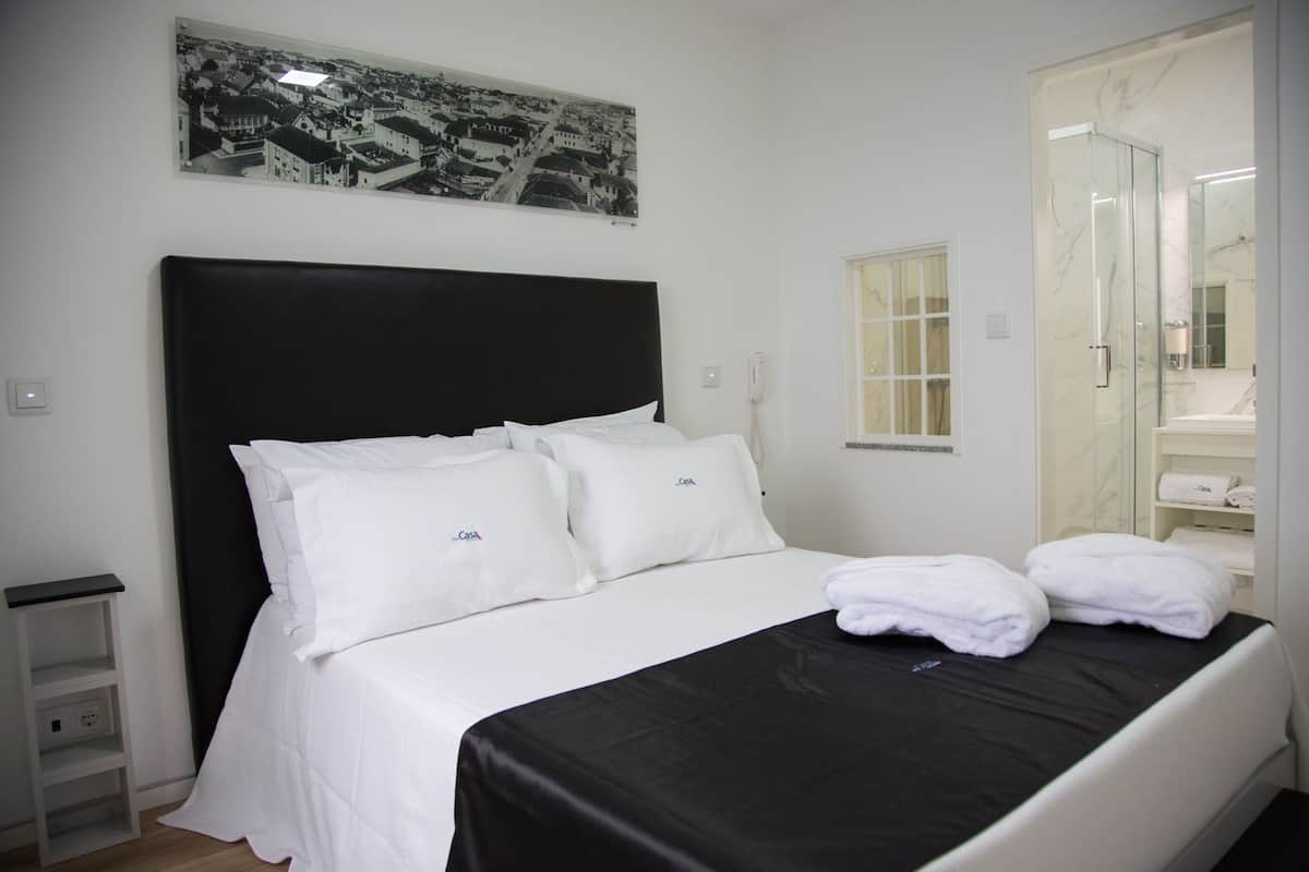 Habitación de hotel con una cama grande, ropa de cama blanca, cabecera negra, un cuadro enmarcado con una vista urbana y una mesita de noche pequeña. Incluye un baño con mampara de vidrio.
