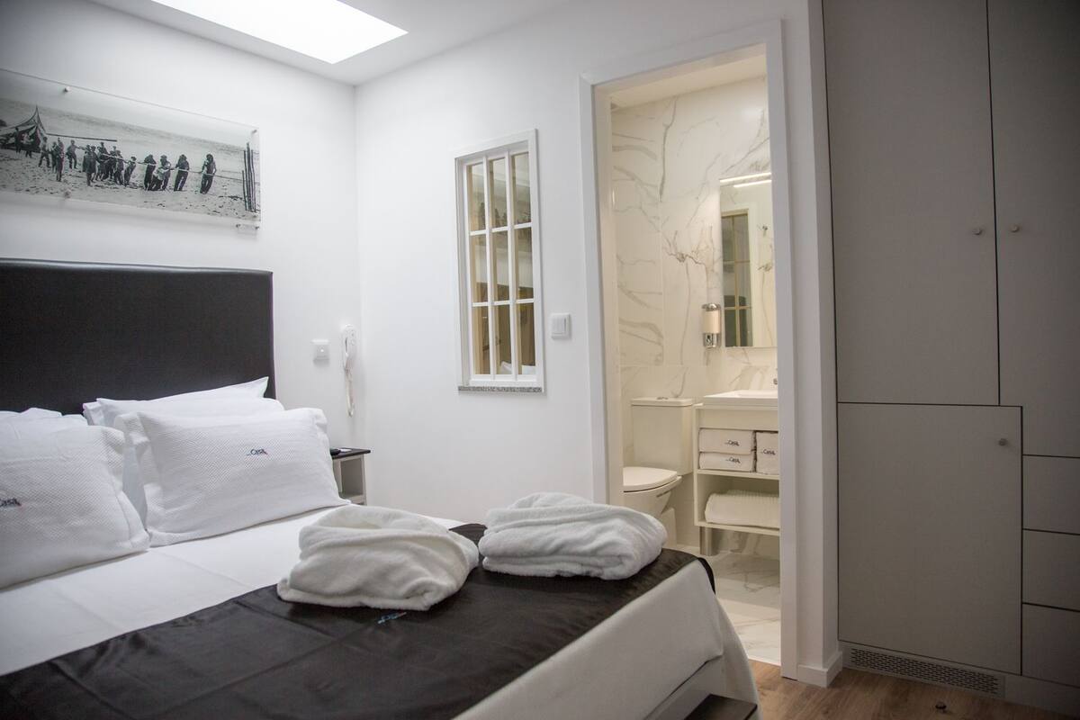 Un dormitorio con cama, mesita de noche, teléfono, ventana y baño visible a través de una puerta abierta.