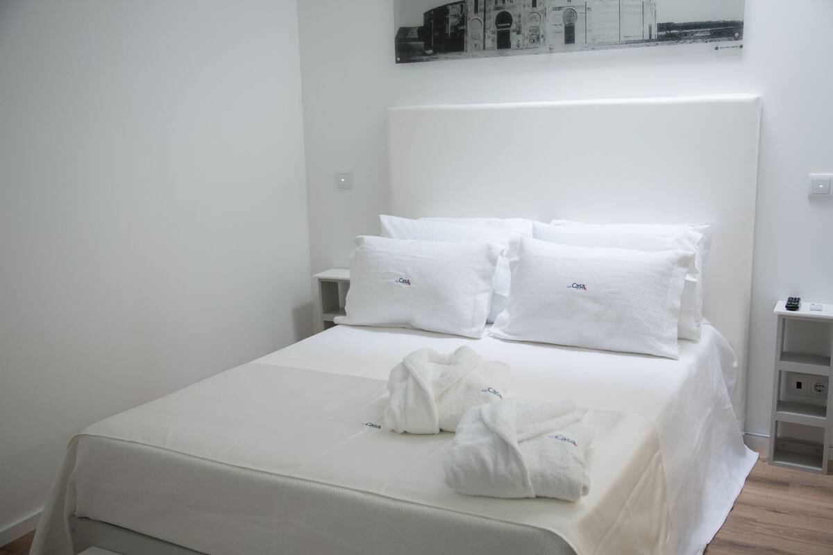 Una cama bien hecha con sábanas y almohadas blancas, una mesita de noche con un control remoto y un cuadro colgado en la pared.