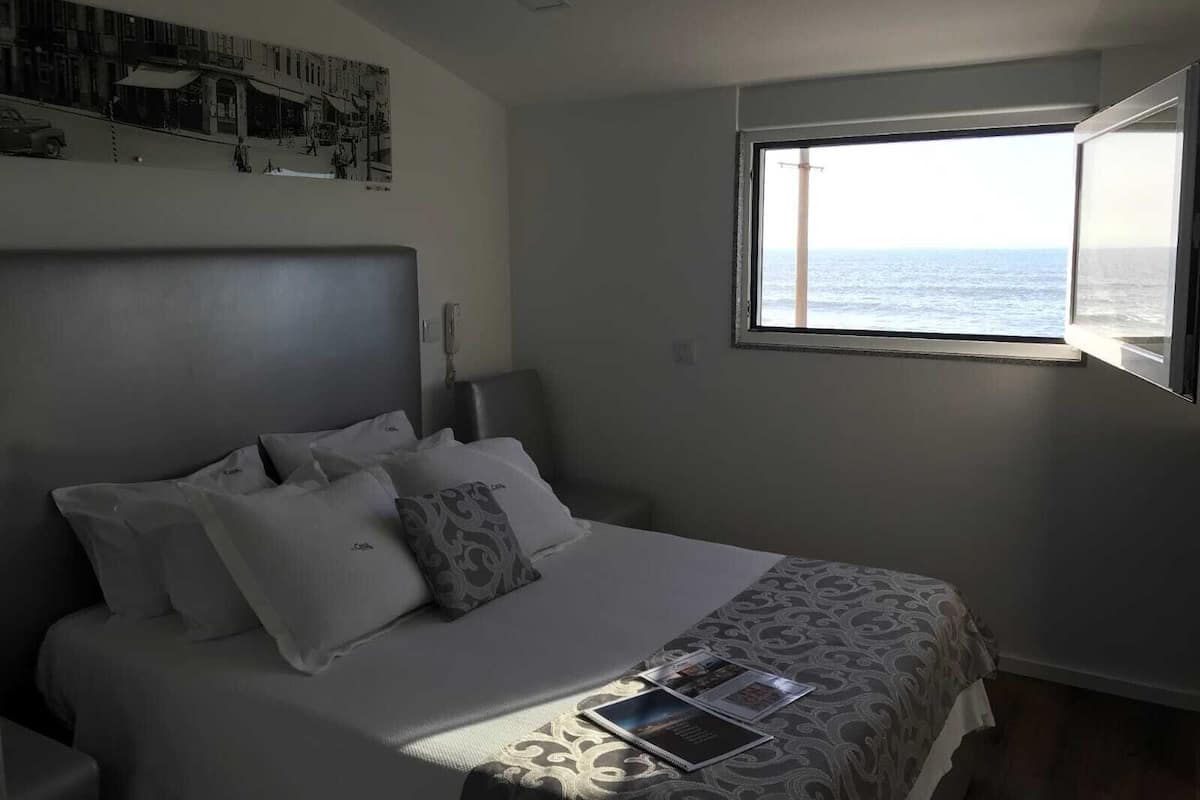 Un dormitorio con cama, almohadas, una mesita de noche, un ventanal con vista al mar y una obra de arte colgada en la pared.