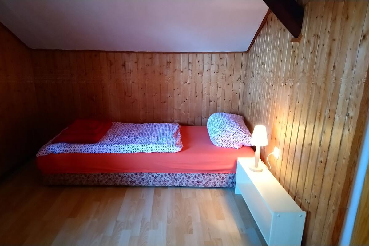 Ein kleiner, gemütlicher Raum mit Holzboden und -wänden, einem Einzelbett mit roter Matratze und gemusterten Kissen sowie einem Nachttisch mit Lampe.